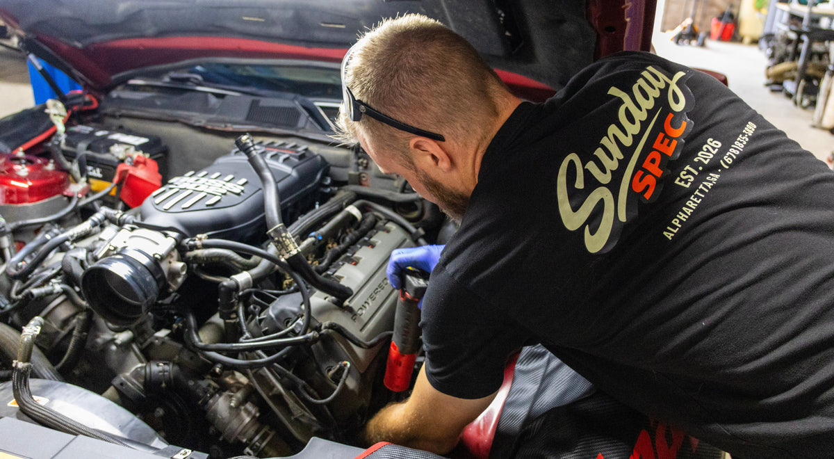 A man in a black t-shirt with 'Sunday SPEC' branding leans over an open car hood, working on the engine in a garage setting.