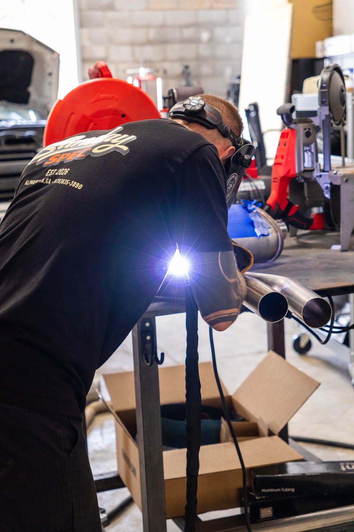 A person in a black t-shirt and welding helmet welding metal pipes on a workbench in a workshop, with a bright welding arc visible.