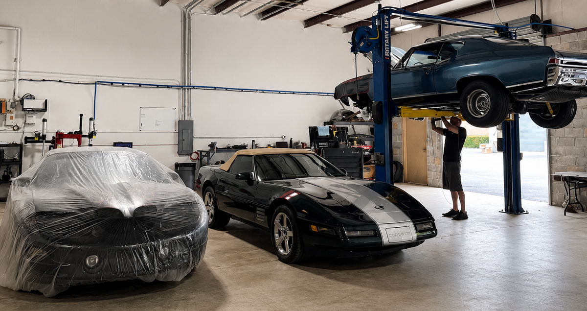 A black sports car with a silver center stripe is parked in a garage next to a car covered in clear plastic. Another dark blue car is elevated on a blue hydraulic lift, and a man stands underneath it.